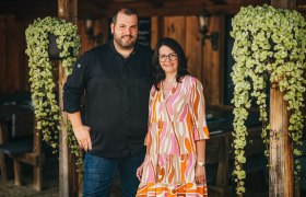 A man and a woman stand smiling next to each other in a rustic room with wooden walls and hanging plants.