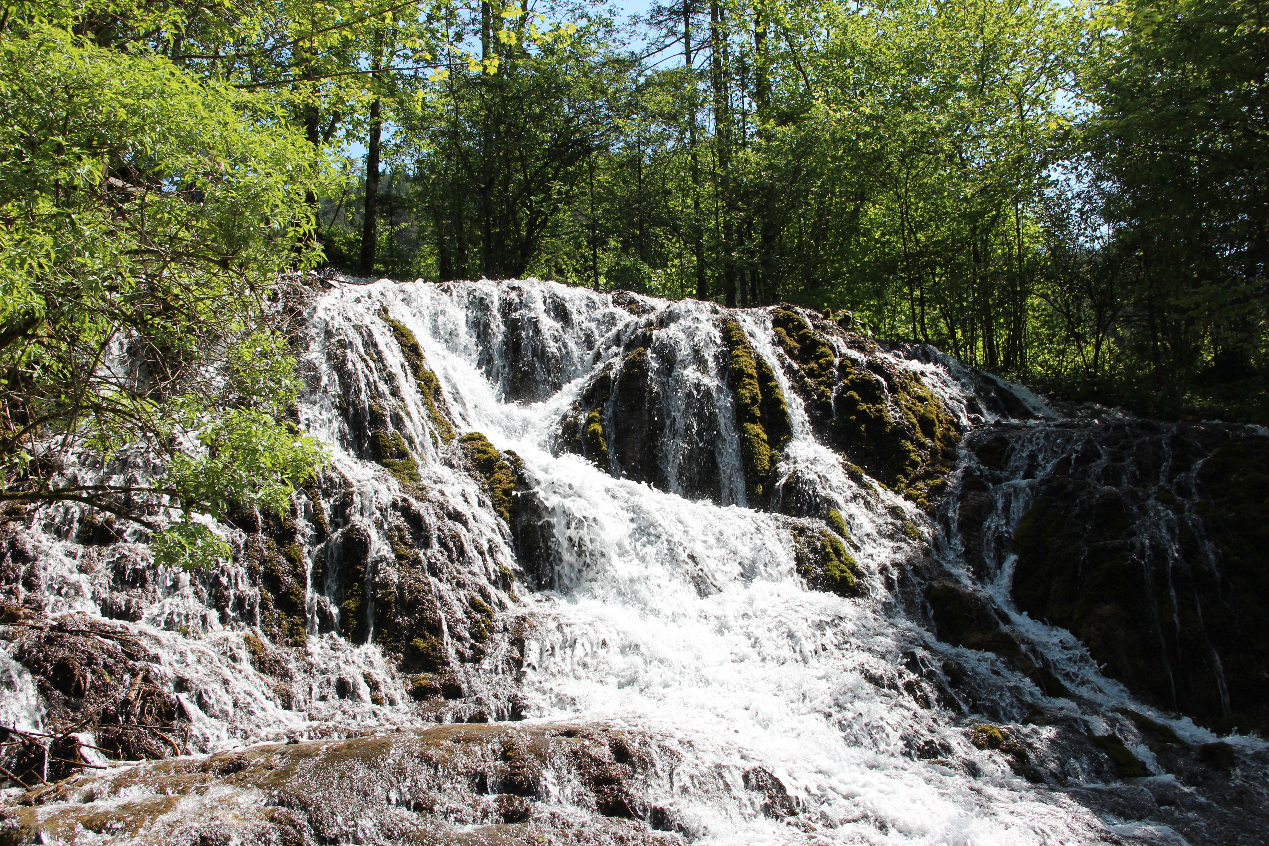 A waterfall flows over moss-covered rocks, surrounded by green trees.