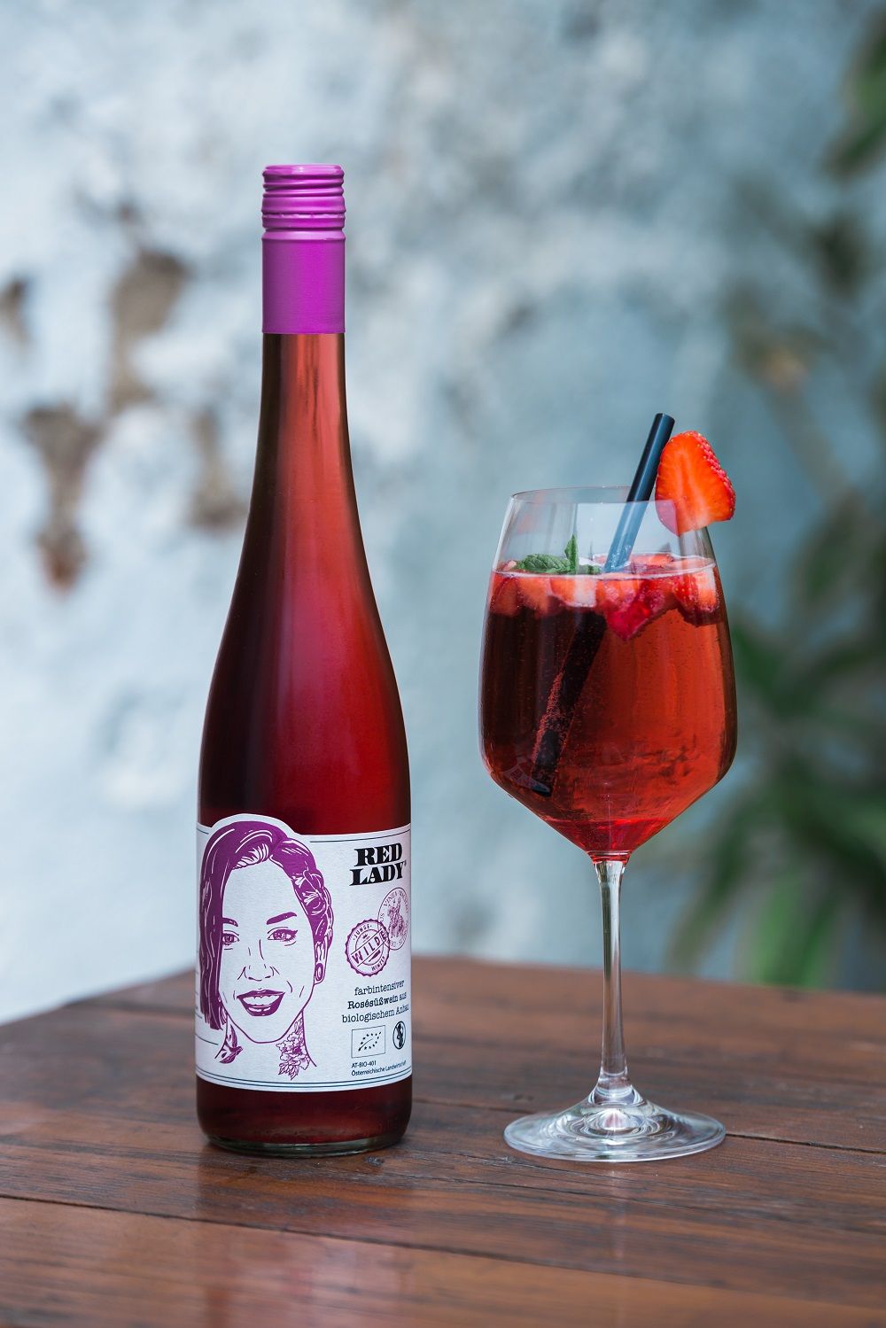 A bottle of red wine with the label 'Red Lady' and a wine glass with a red drink and strawberry on a wooden table.