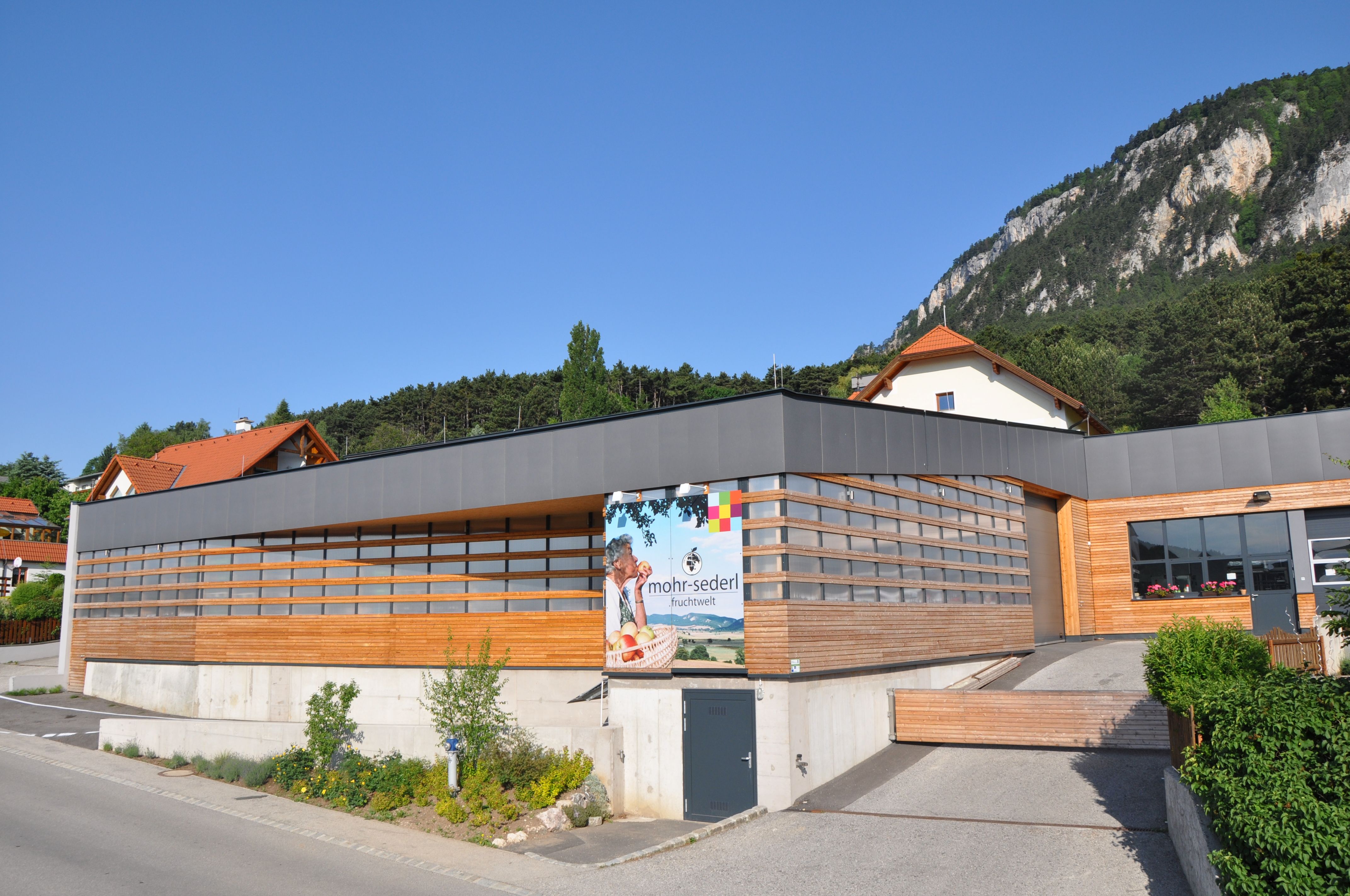 Modern building with wooden cladding and advertising sign for Mohr-Sederl Fruchtwelt, surrounded by green countryside and mountains.
