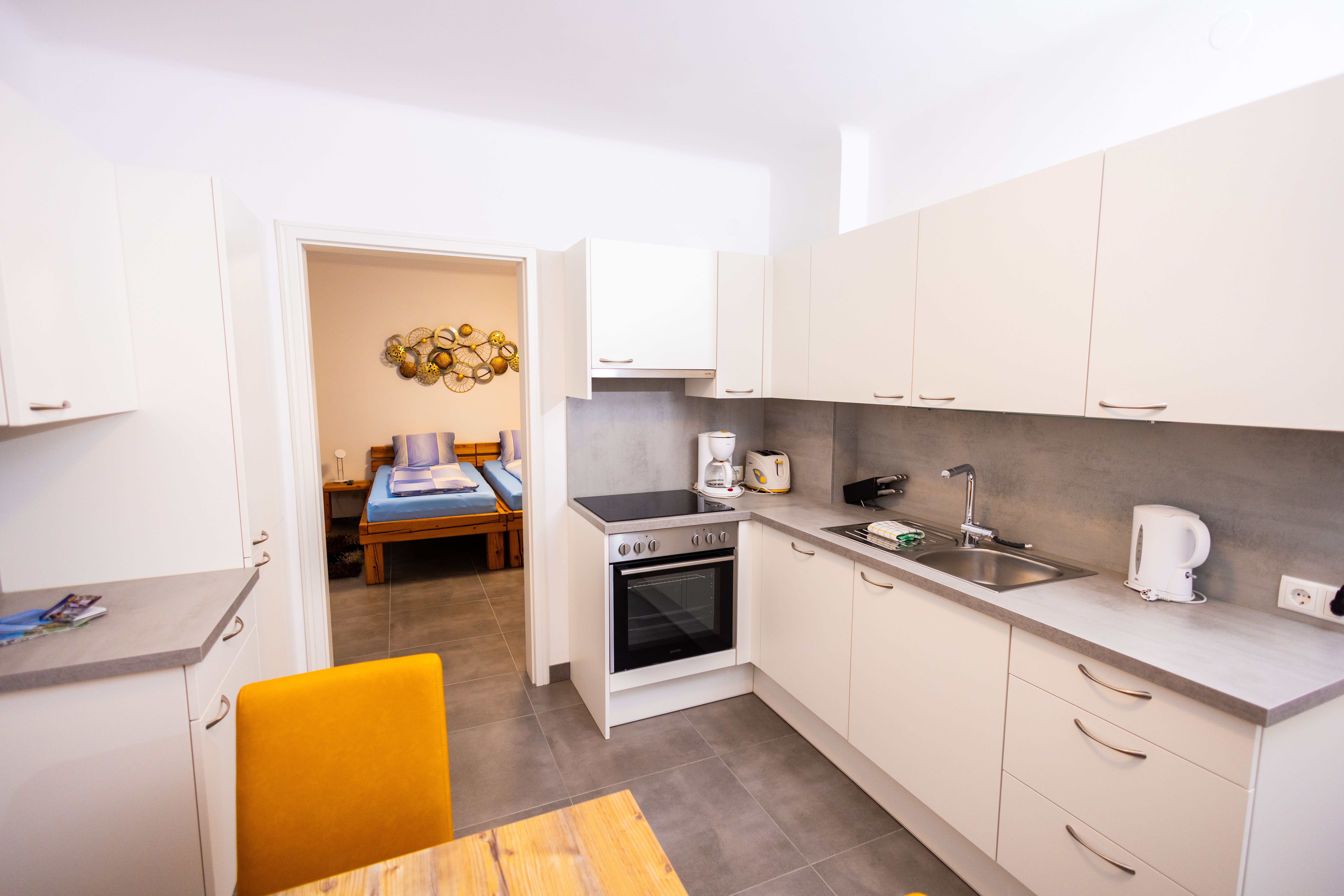 Modern kitchen with white cupboards, stove, sink and view into a bedroom with double bed.