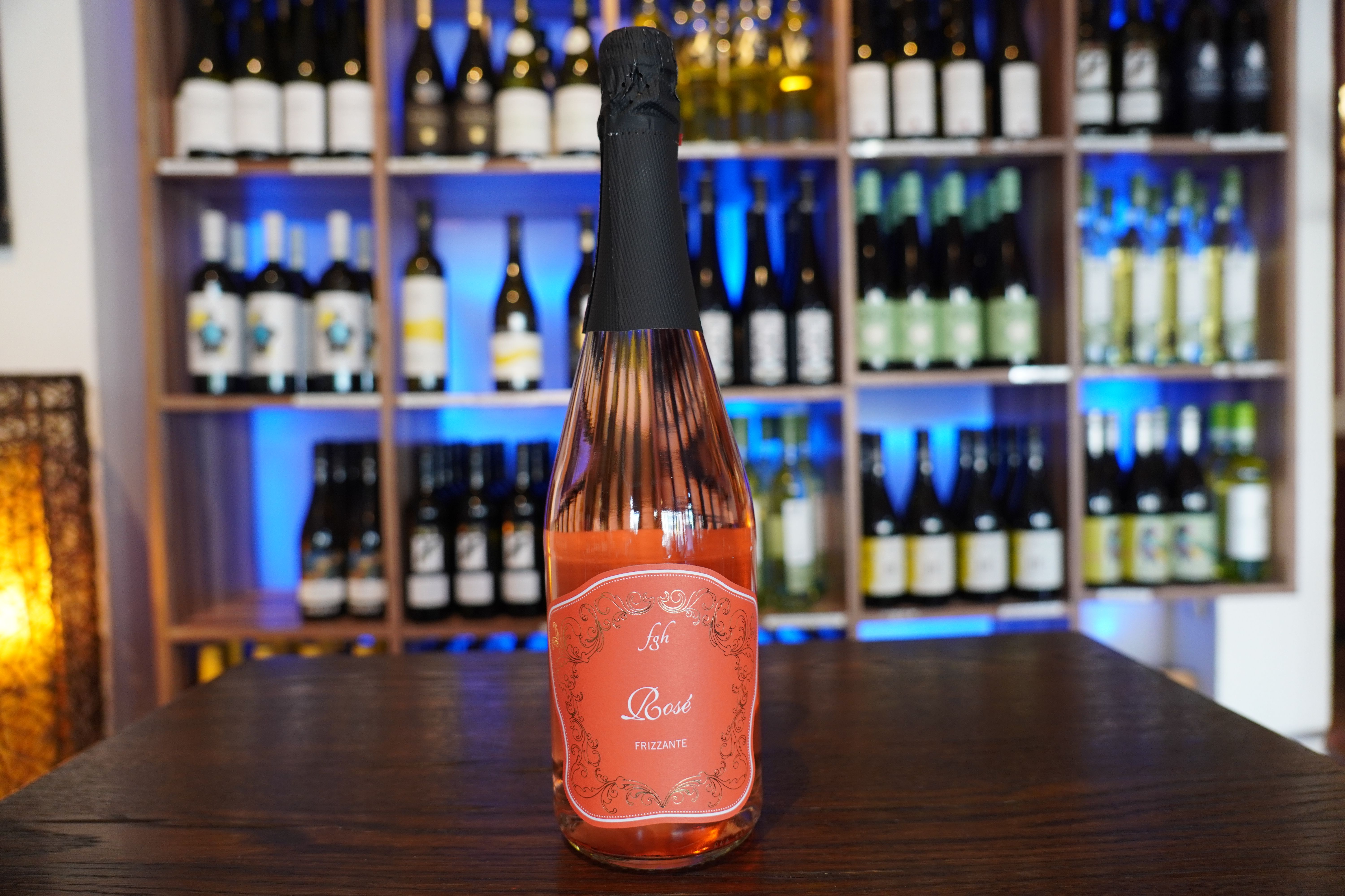 A bottle of rosé wine stands on a table in front of a wine rack.