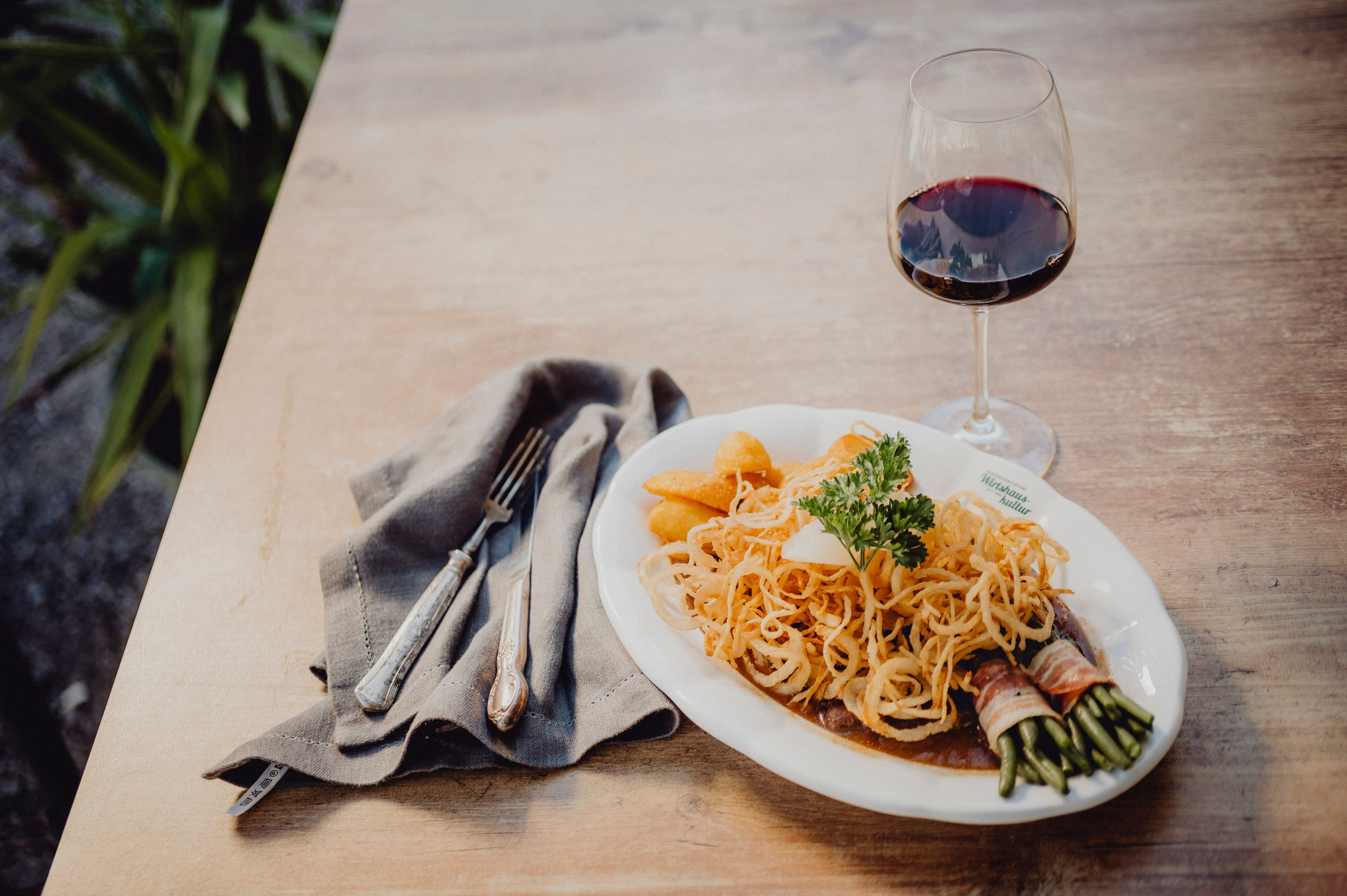 A plate of roast onions, potatoes and bacon beans, next to a glass of red wine on a wooden table.