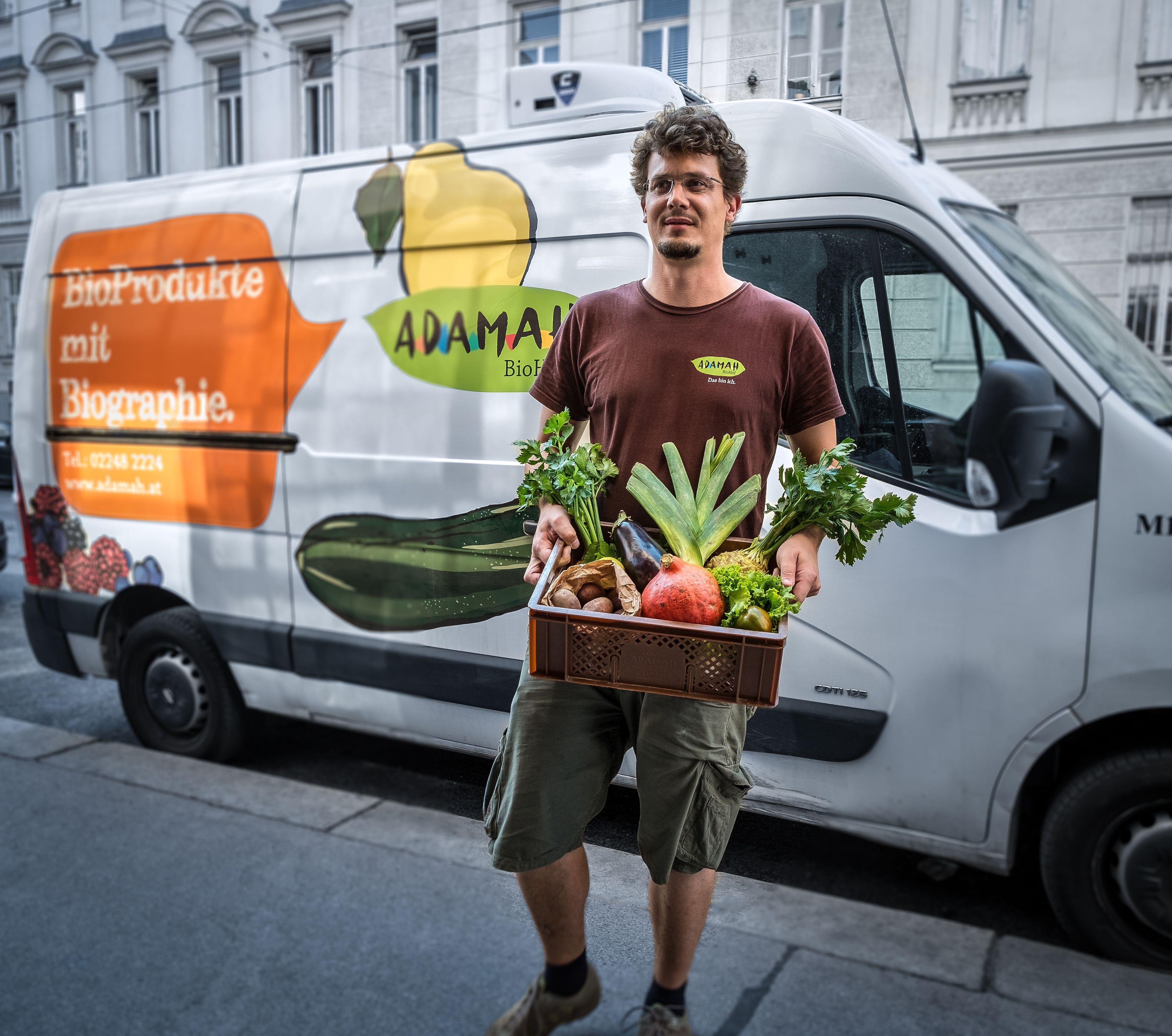 A man holds a box of fresh fruit and vegetables in front of a delivery van with the ADAMAH logo.