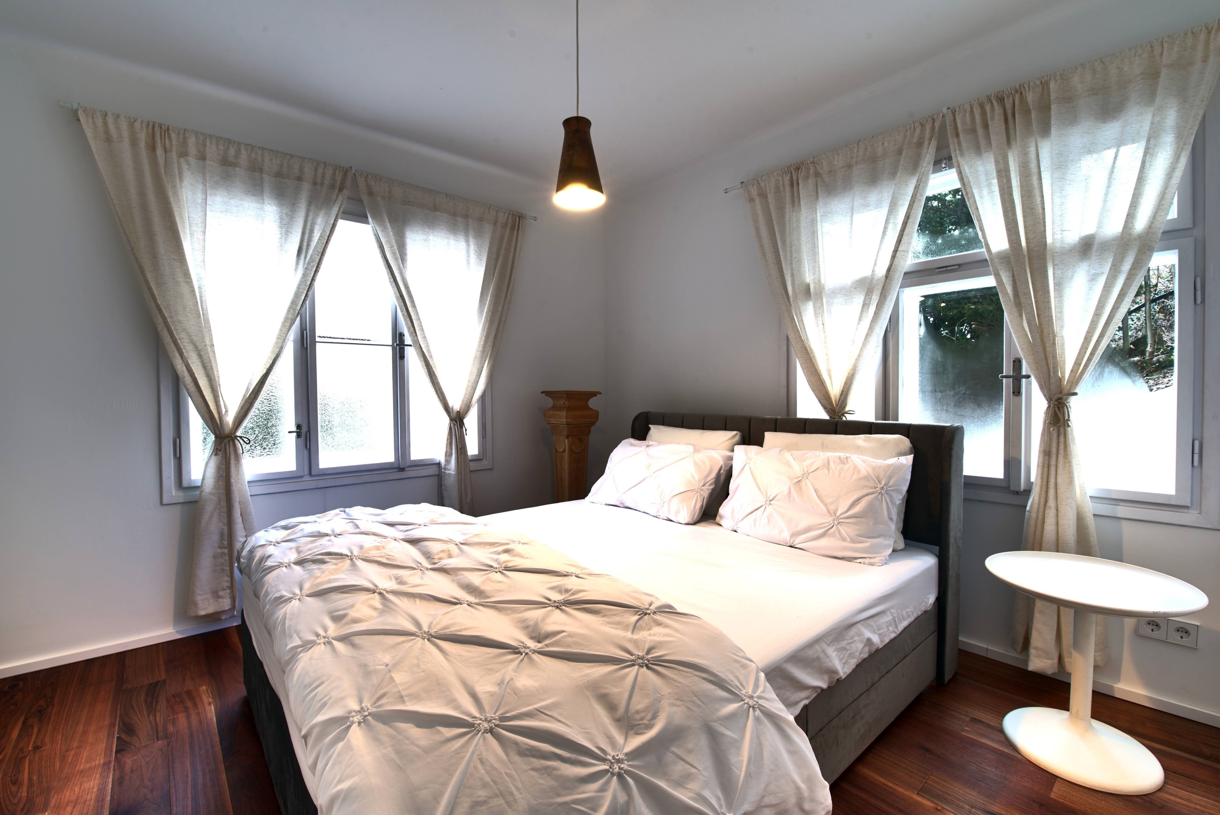 A cozy bedroom with a large bed, white curtains and a small round table.