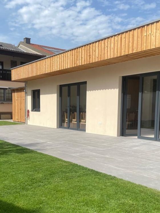 Modern building exterior with wooden cladding and large windows, surrounded by a terrace and lawn.