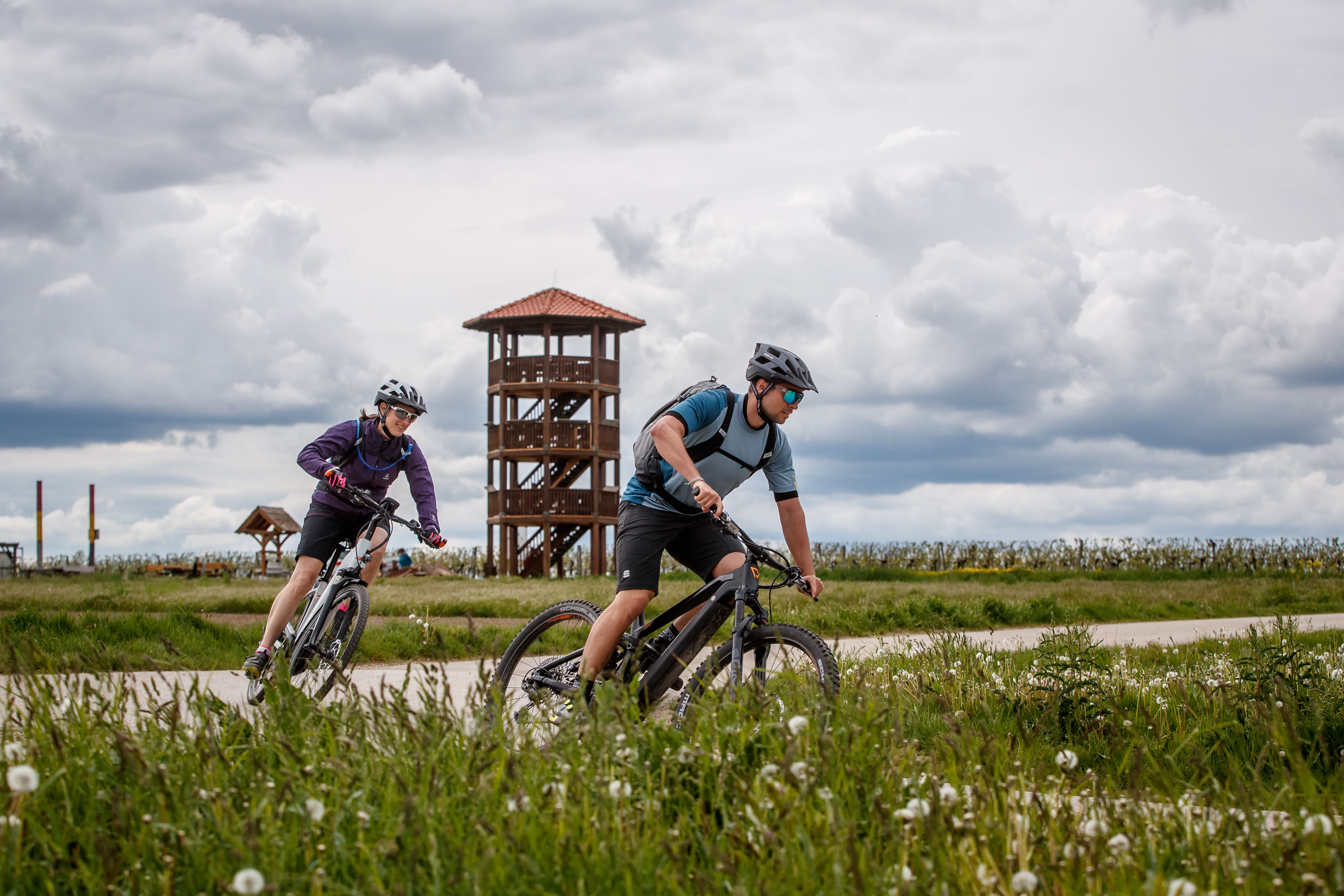 Two cyclists ride along a path in front of a lookout tower, surrounded by a green landscape and a cloudy sky.