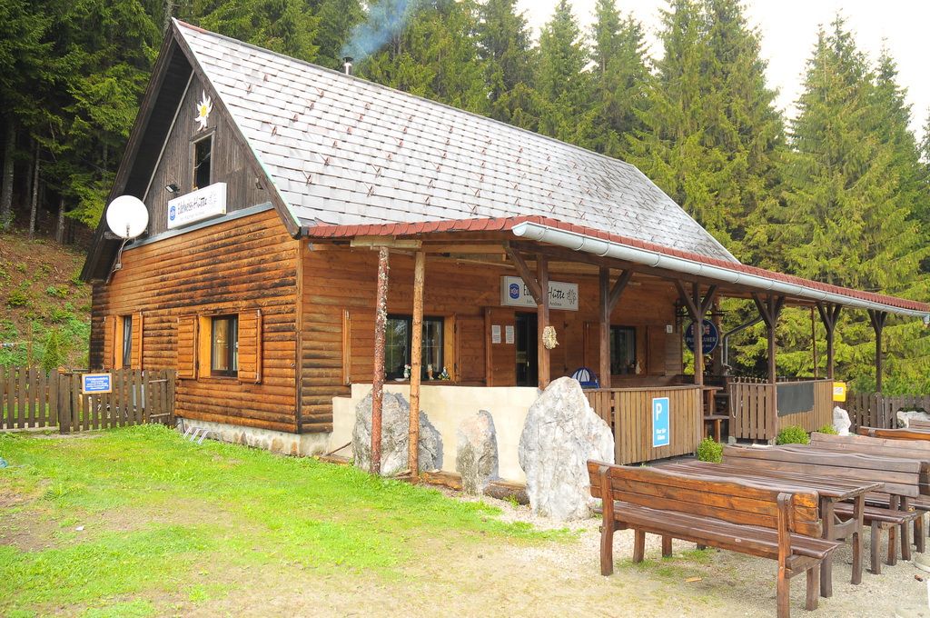 A wooden hut in the forest with tables and benches in front of it.