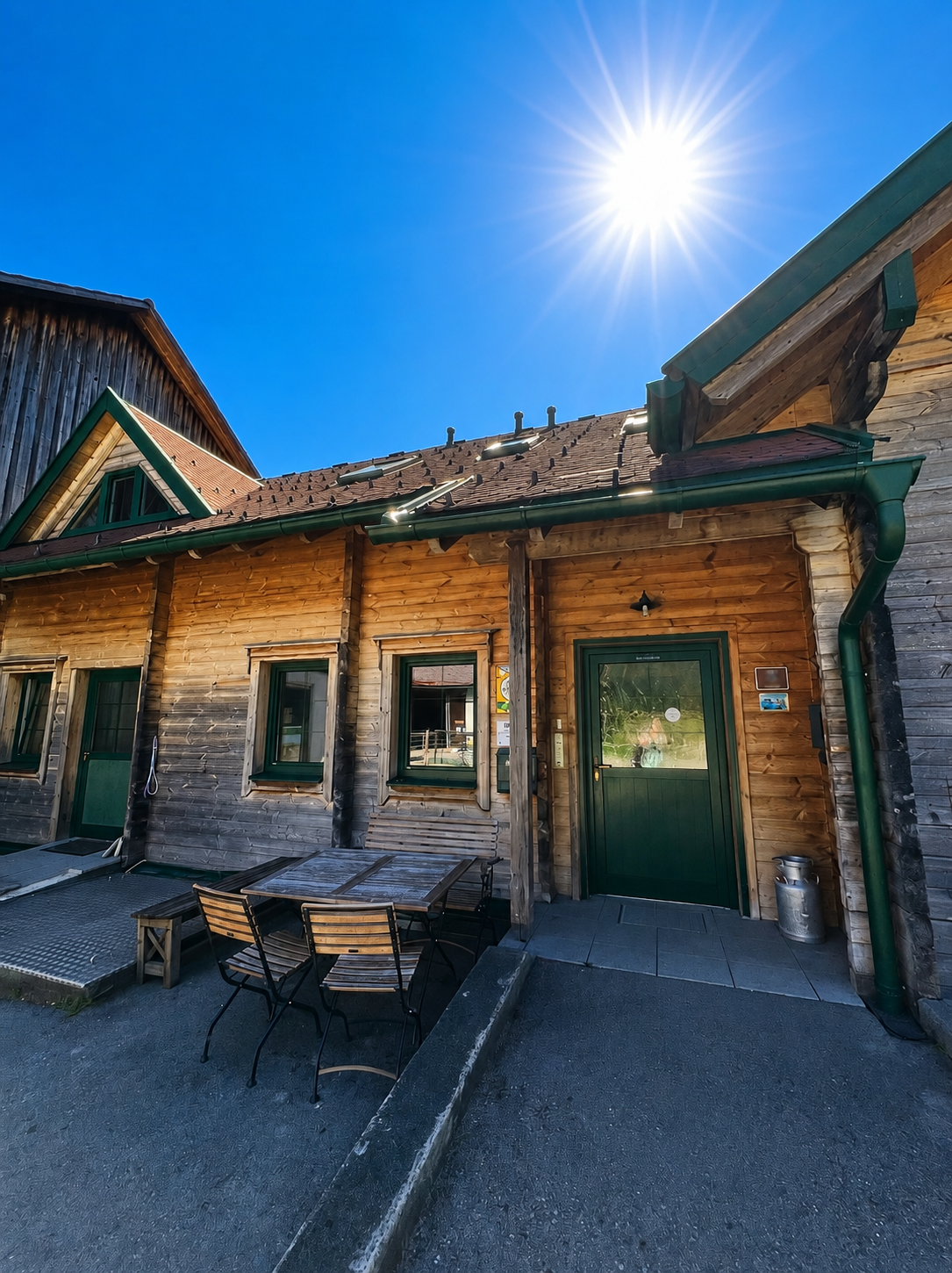 A wooden building with a green roof and a table with chairs in the foreground, under a bright sun.