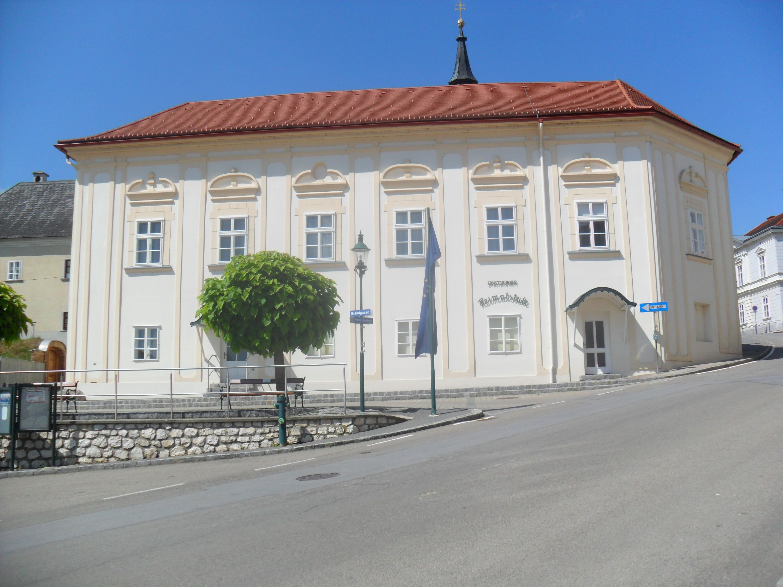 Historic building with a red roof and white walls, surrounded by trees and a road.