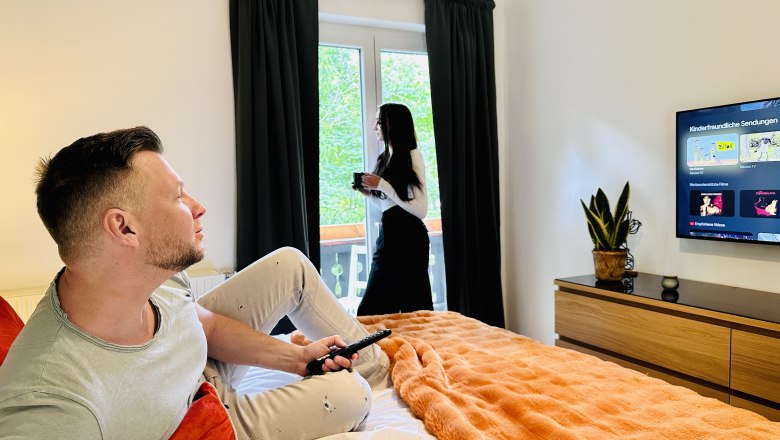 A man sits on a bed and watches television while a woman stands at the window drinking coffee.