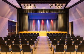 An empty event hall with rows of chairs and a stage with curtains and colorful lighting.