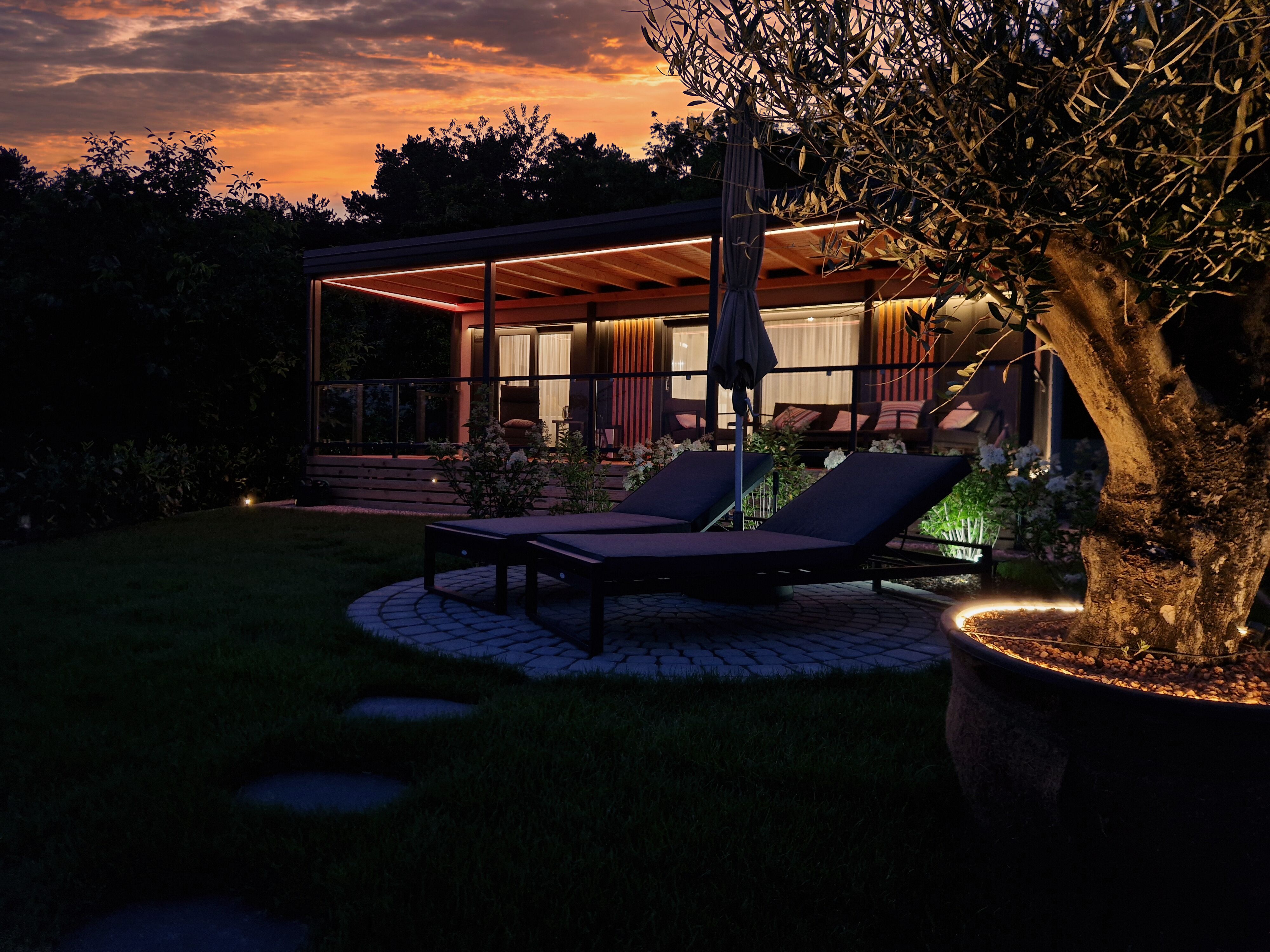 Exterior view of a chalet at sunset with sun loungers and illuminated tree in the foreground.