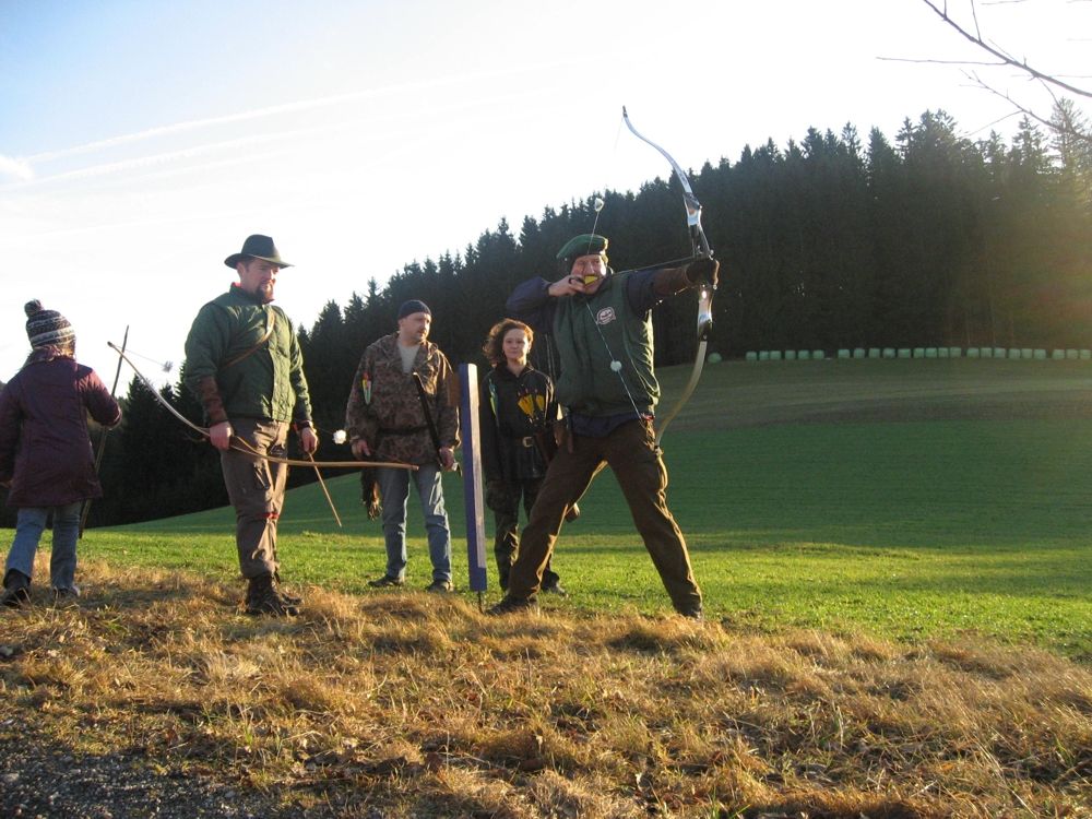 Group of people doing archery in a meadow in front of a forest.