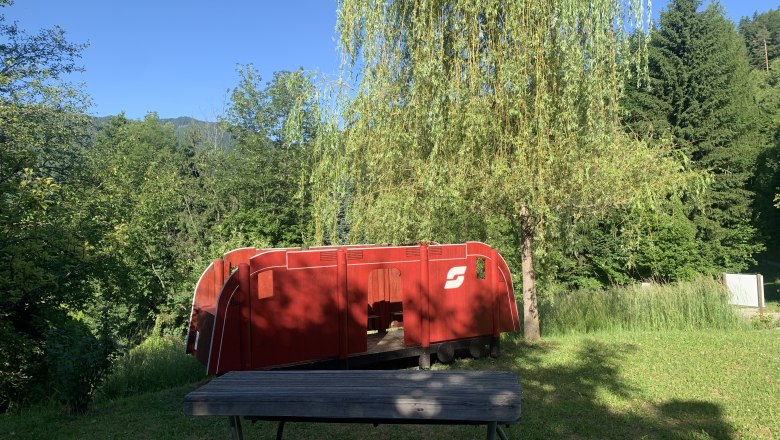 Red toy locomotive outdoors next to a tree and a wooden bench.