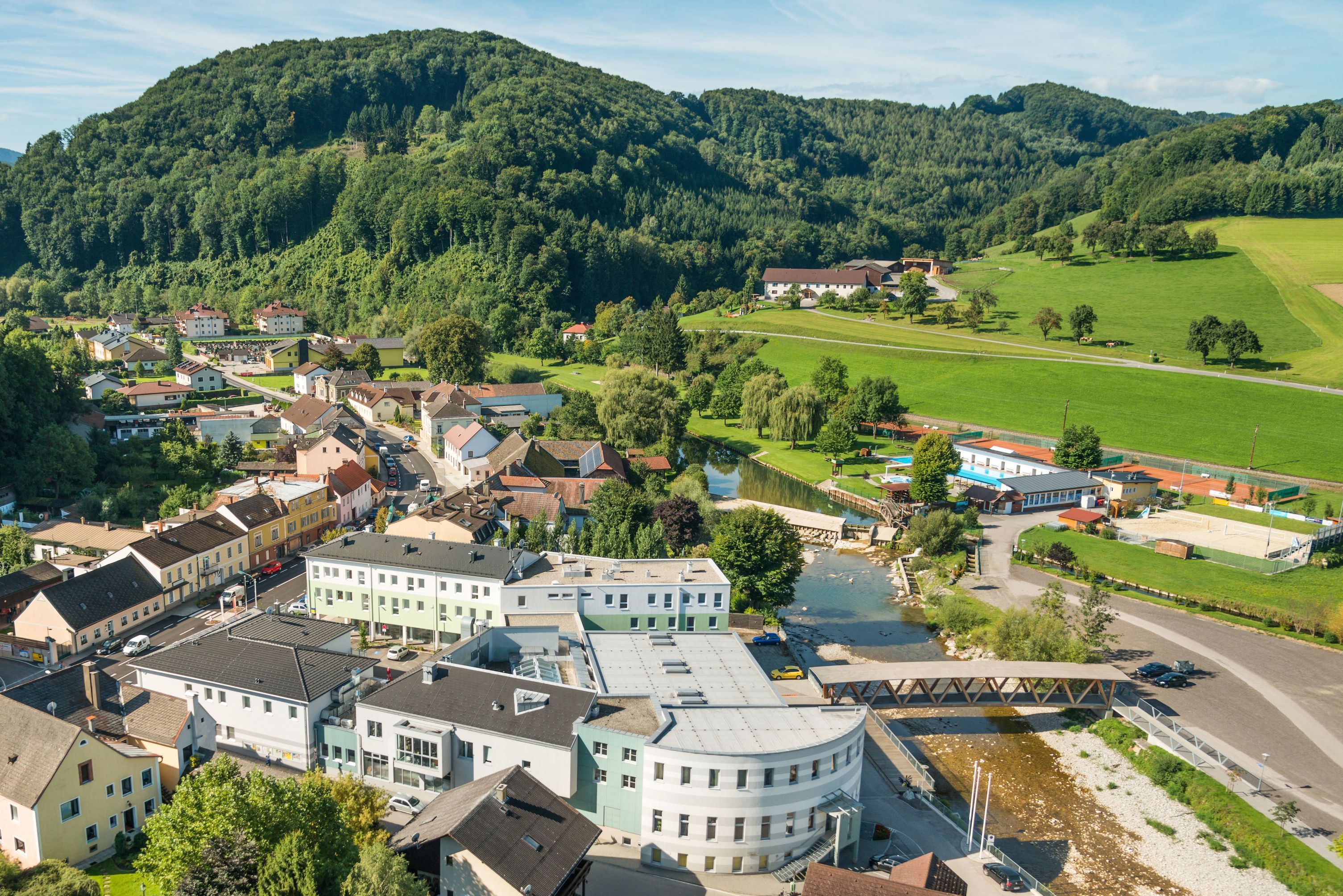 Aerial view of a small village with river, bridge and surrounding hills.
