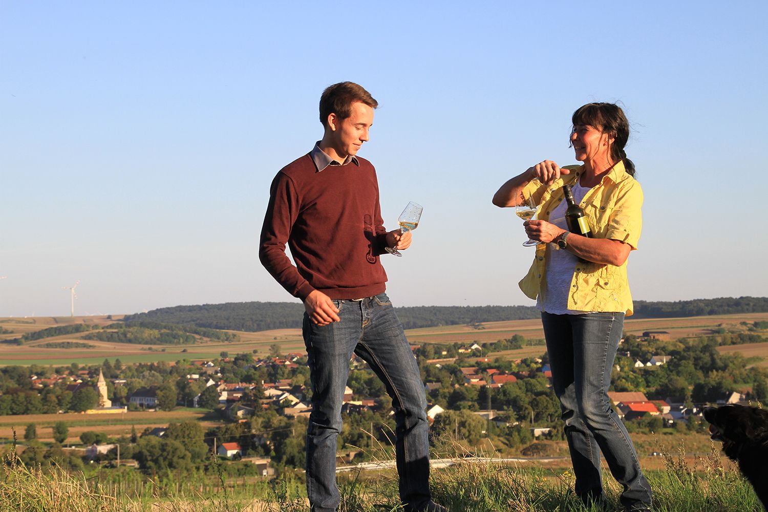 Two people stand on a hill with wine glasses in their hands, in the background a rural landscape with wind turbines.