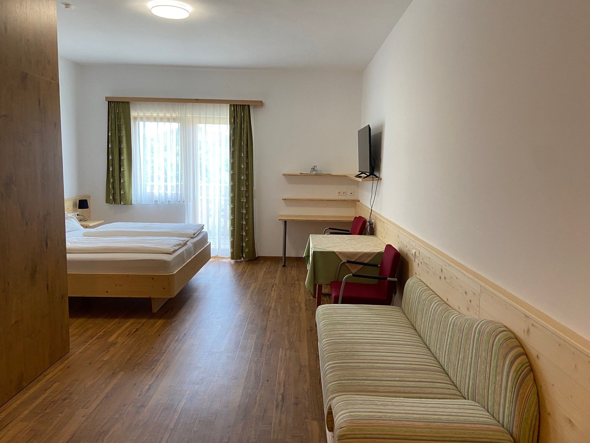 A modern hotel room with a double bed, sofa, table and chairs. A television hangs on the wall. Curtains on one window let in daylight.