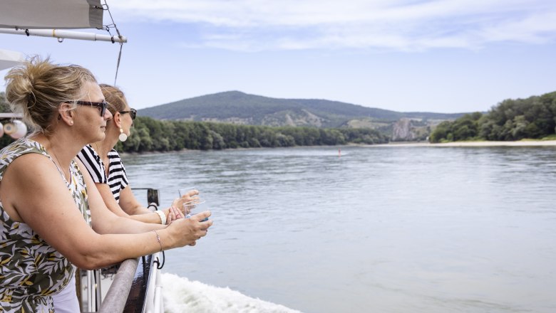 Two women stand at the railing of a boat on the Danube and enjoy the view of the surrounding landscape.