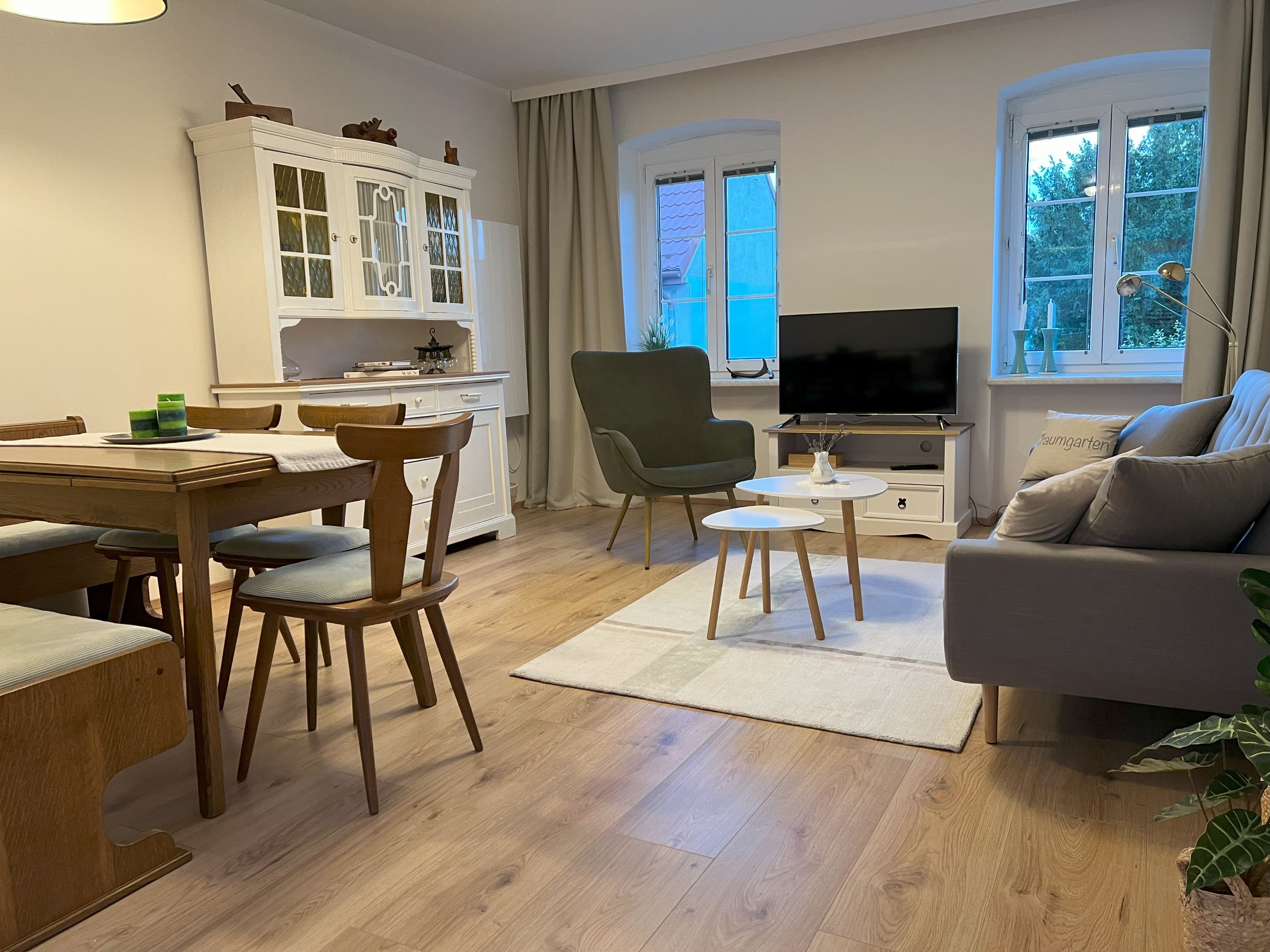 Cozy living room with wooden furniture, sofa, armchair, dining table and TV. Light-colored walls and wooden floor.