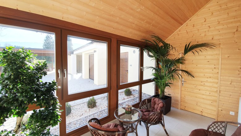 Interior view of a veranda with wooden walls, large windows, plants and rattan furniture.