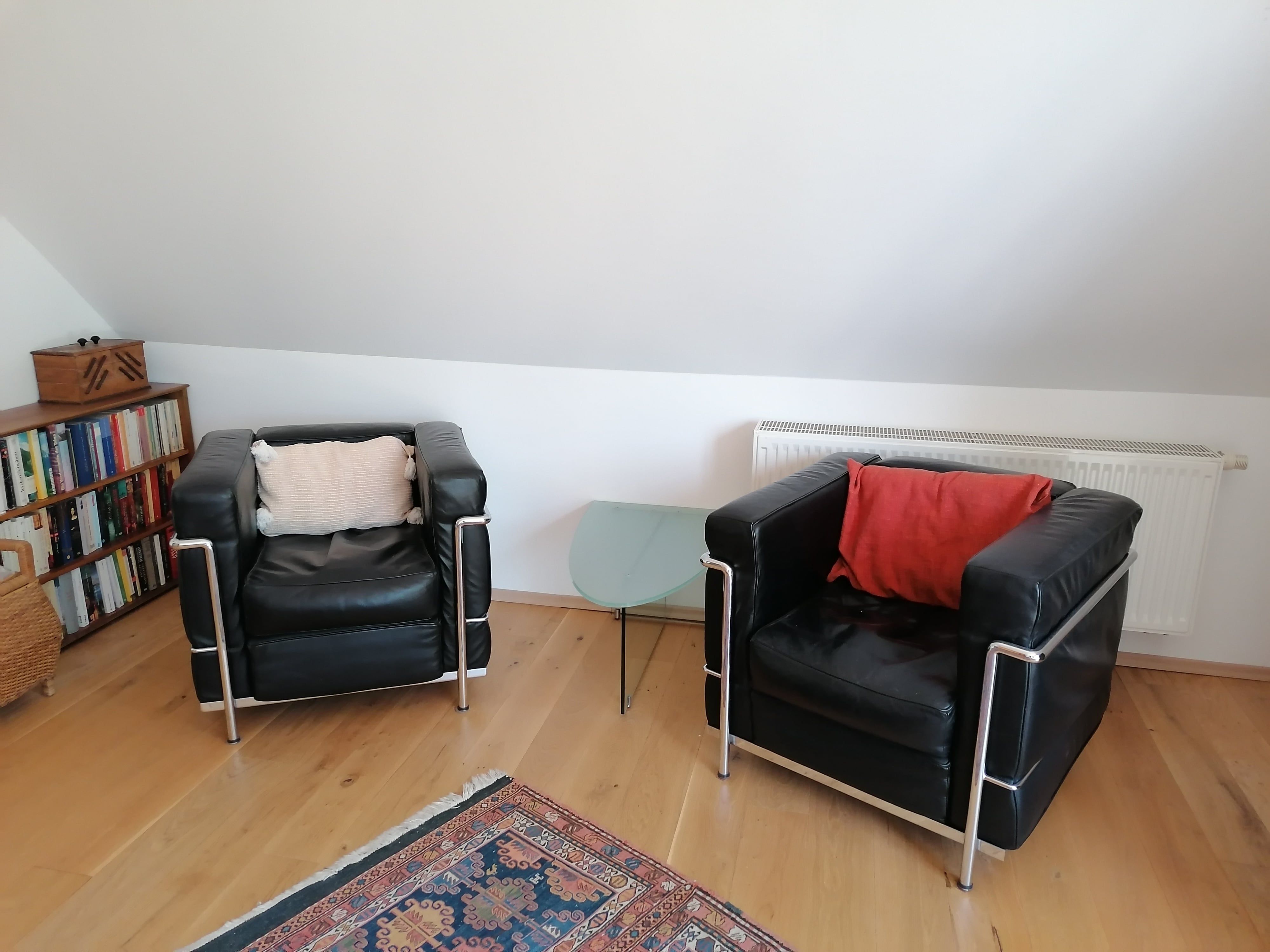 Two black leather armchairs with cushions, a glass side table, bookshelf and carpet in a living area with a sloping roof.