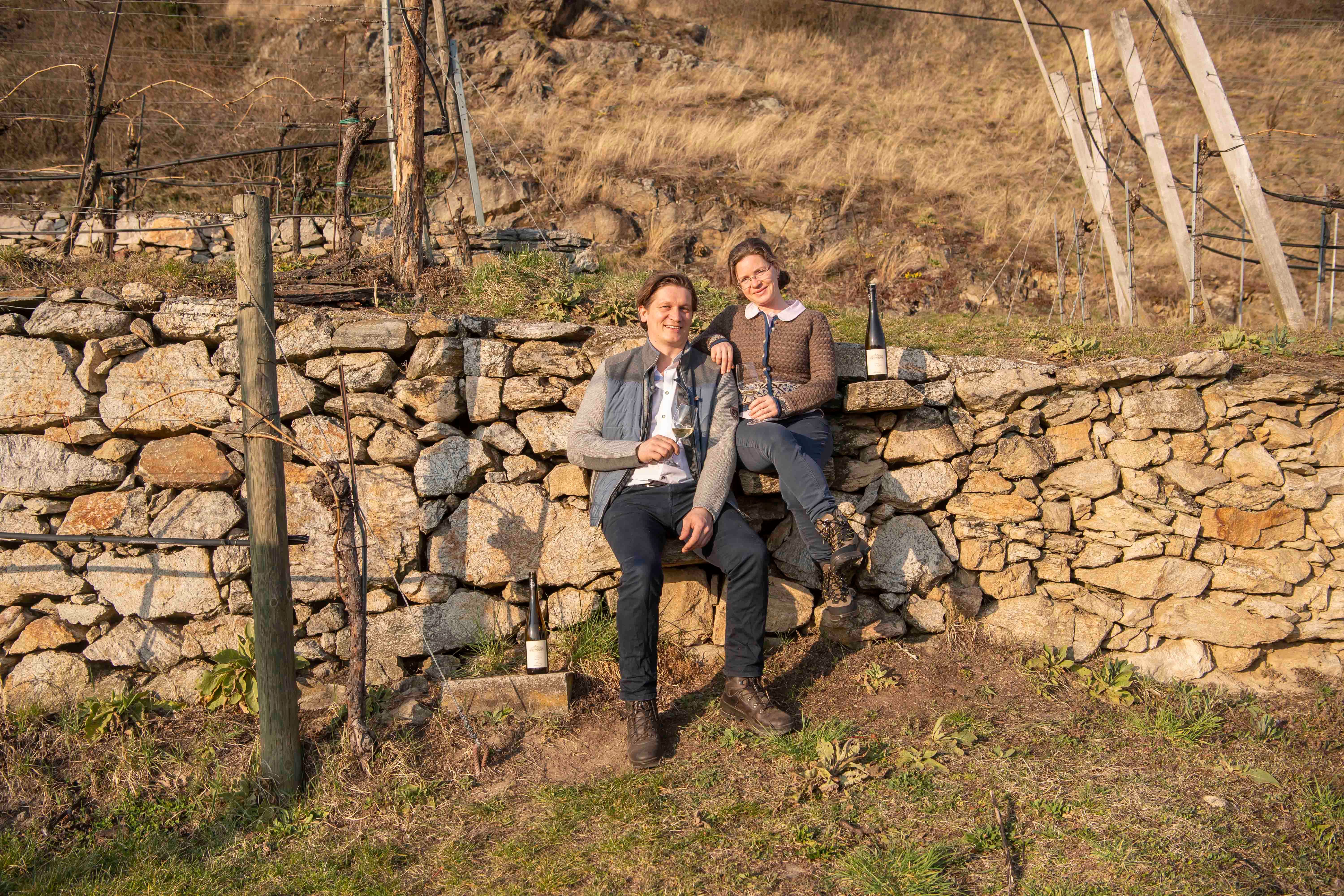 Two people are sitting on a stone wall in a vineyard holding wine glasses.