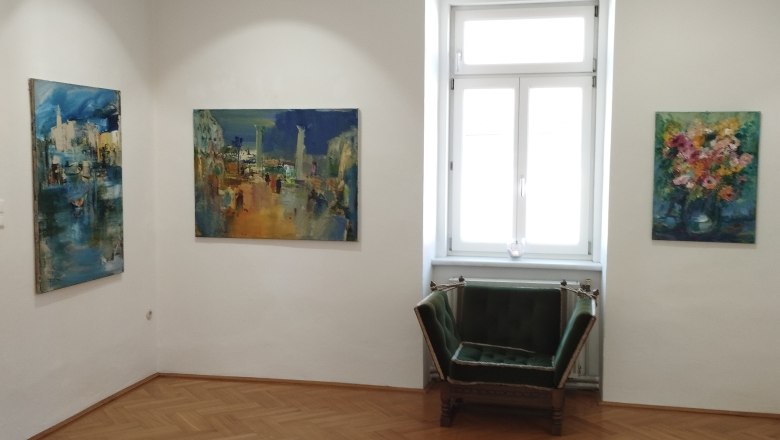 Interior view of a gallery with three paintings on the walls and a green armchair in front of a window.
