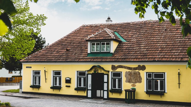Yellow building with red roof tiles and green shutters, surrounded by trees.