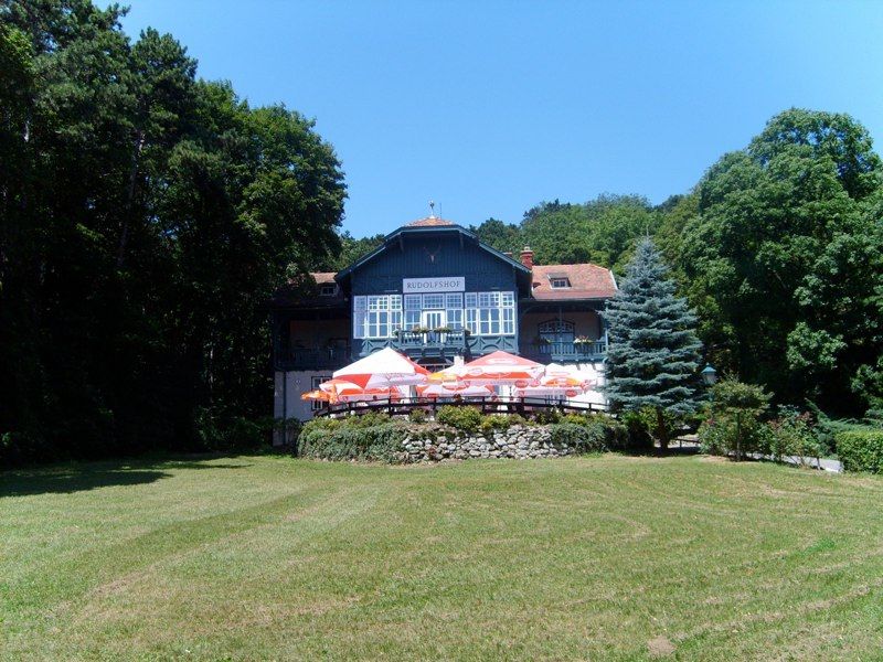 A large house called Rudolfshof with a terrace and sunshades, surrounded by trees and a meadow.