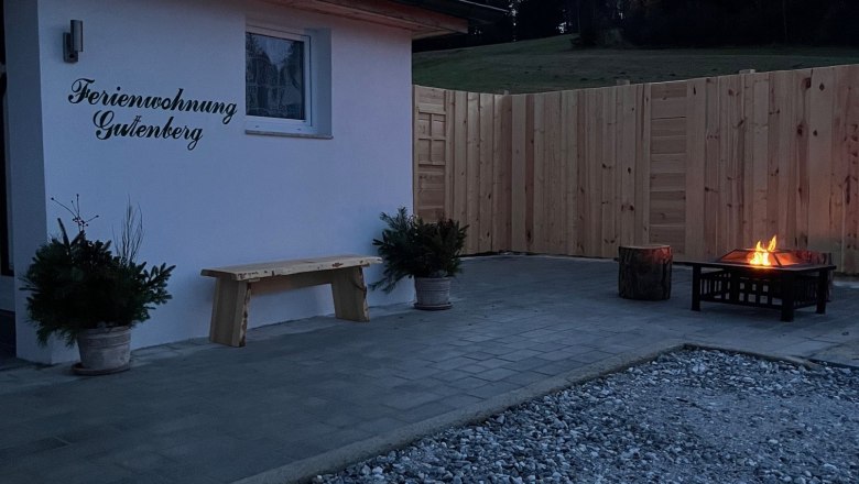 Terrace with fireplace and wooden fence at dusk, next to a building labeled 'Ferienwohnung Gutenberg'.