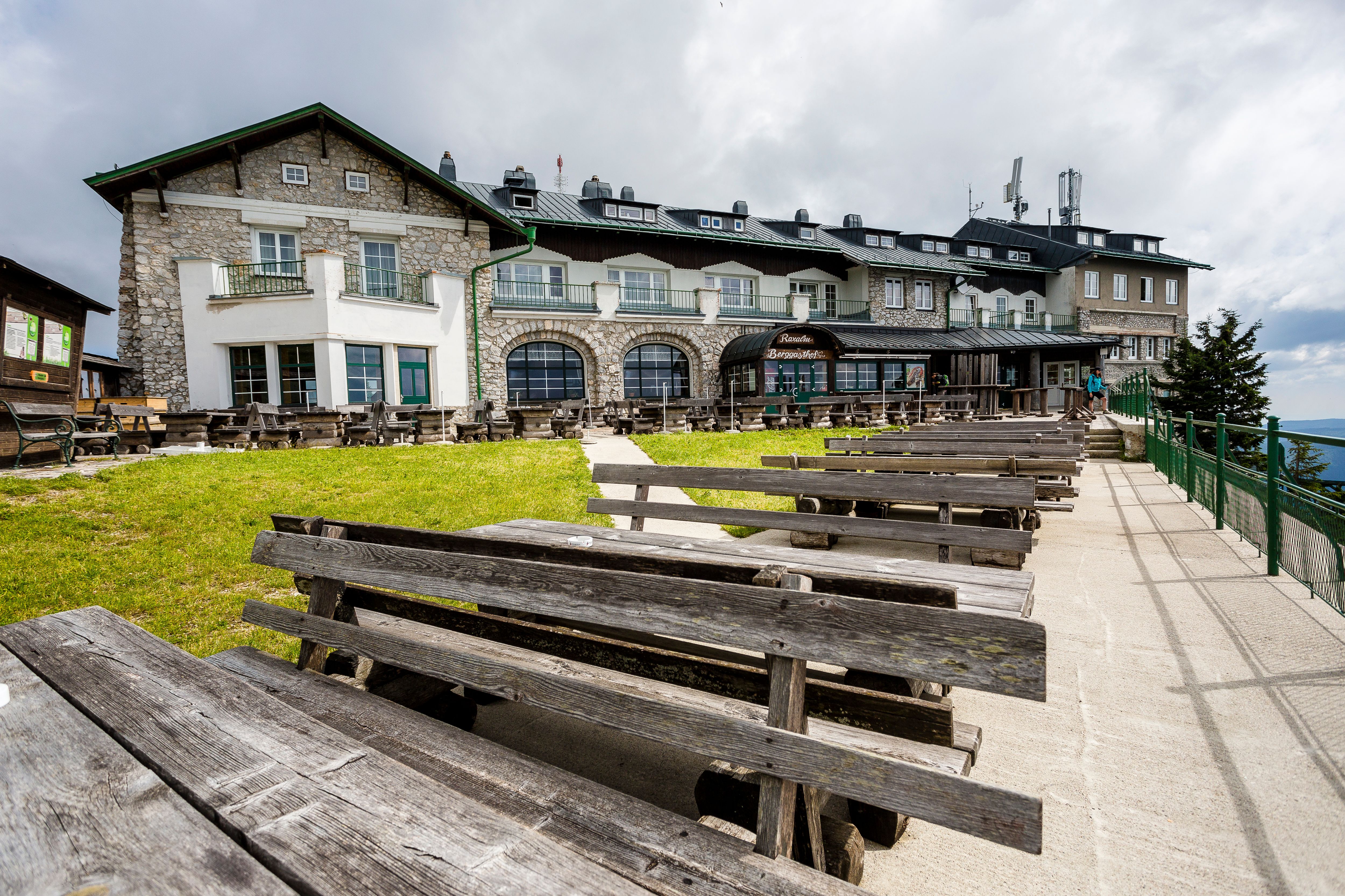 A mountain inn with wooden benches and tables outside, surrounded by green meadows and a cloudy sky.