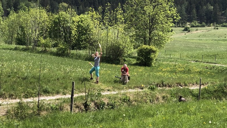 Two people in a meadow, one shooting with a bow, the other sitting. Trees and hills in the background.