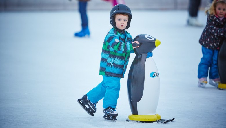 A child wearing a helmet and colorful jacket holds on to a penguin skating aid on an ice rink.