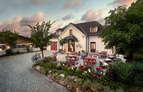 A cozy courtyard with tables, chairs and flower beds in front of a large house at sunset.