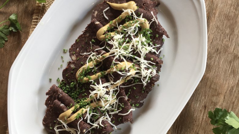 A plate of Blunzenbrot, garnished with mustard, chives and grated cheese, on a wooden table with parsley and a knife next to it.