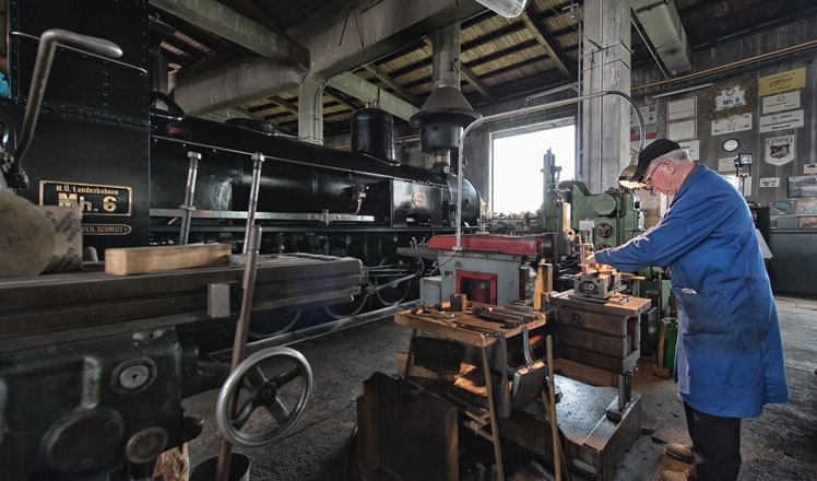 A man works in a workshop on a machine next to an old steam locomotive.