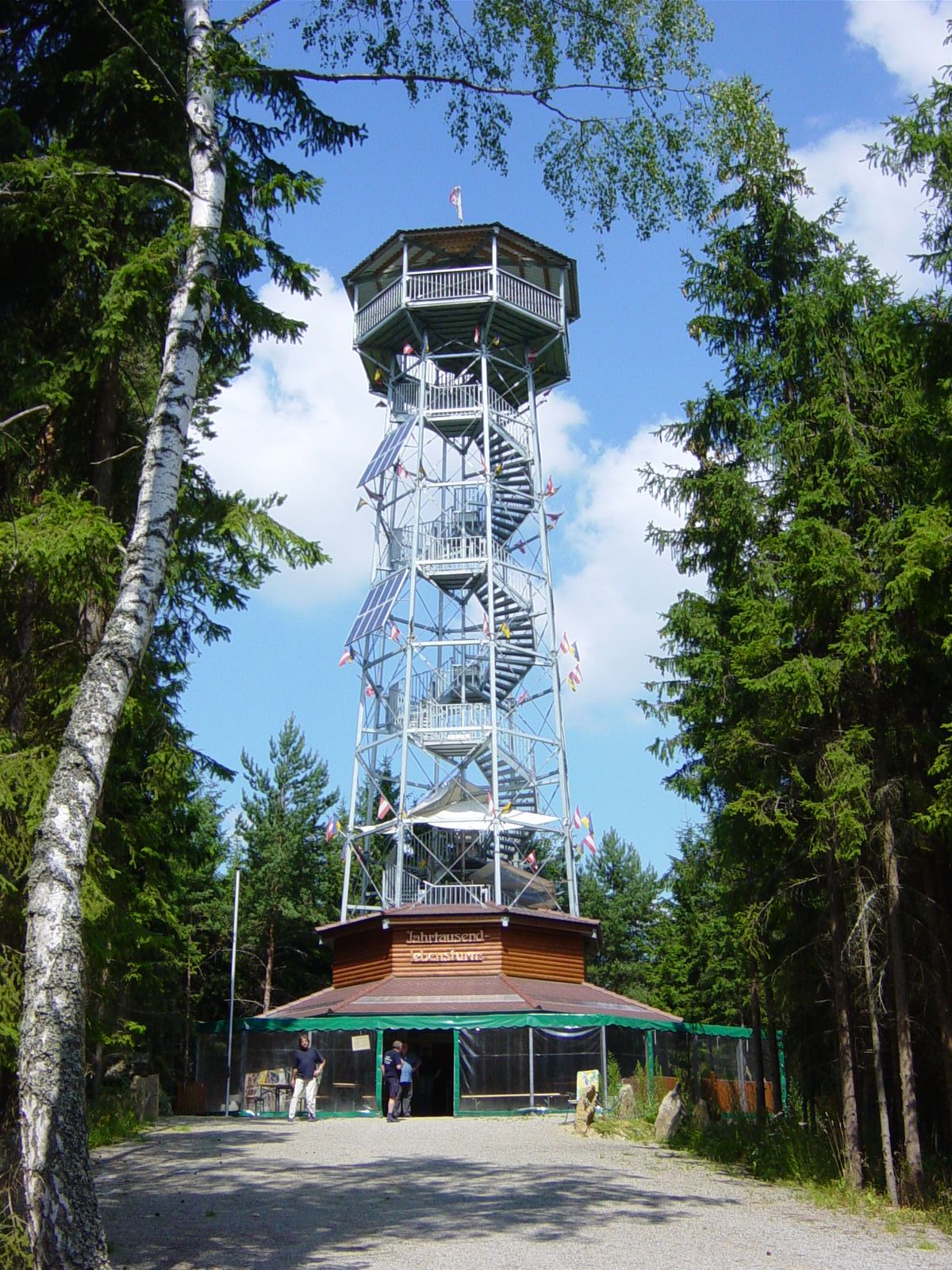 A tall metal observation tower with wooden cladding, surrounded by trees, under a blue sky.