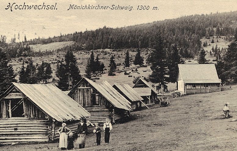 Historical photograph of wooden huts on the Mönichkirchner Schwaig, surrounded by forest and meadows, with people in the foreground.