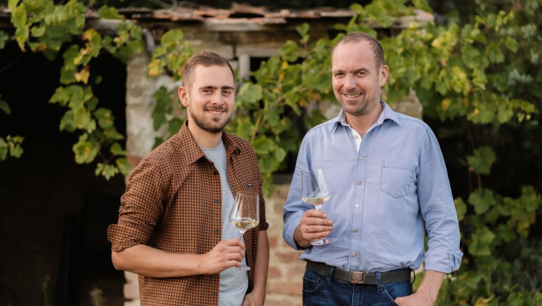 Two men stand smiling with wine glasses in front of a building covered in vine leaves.