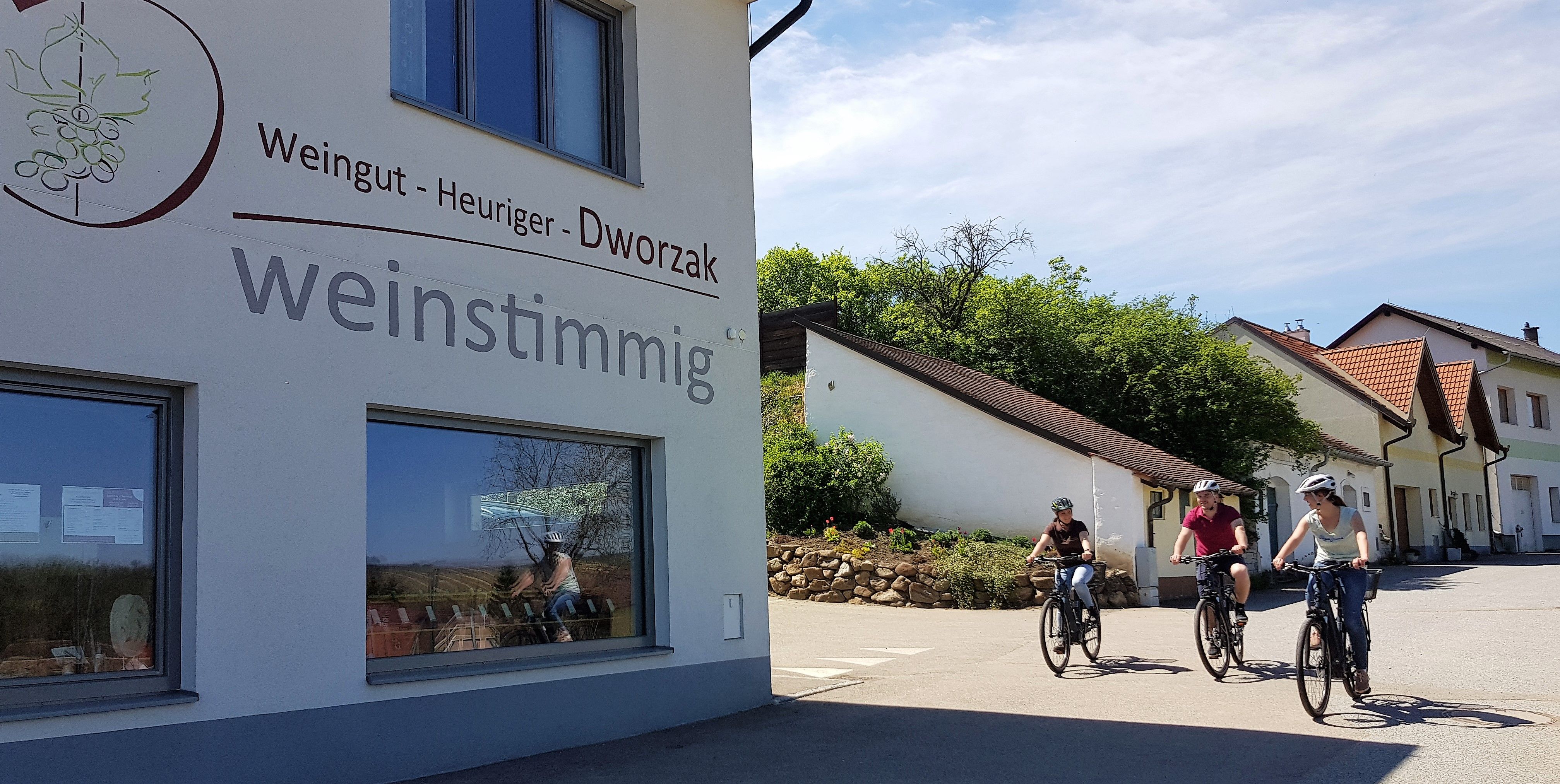 Three people on bicycles ride past a building with the sign 'Weingut - Heuriger - Dworzak weinstimmig'.