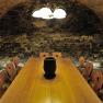 A rustic wine cellar with a long wooden table and chairs, surrounded by stone walls and illuminated by a ceiling lamp.