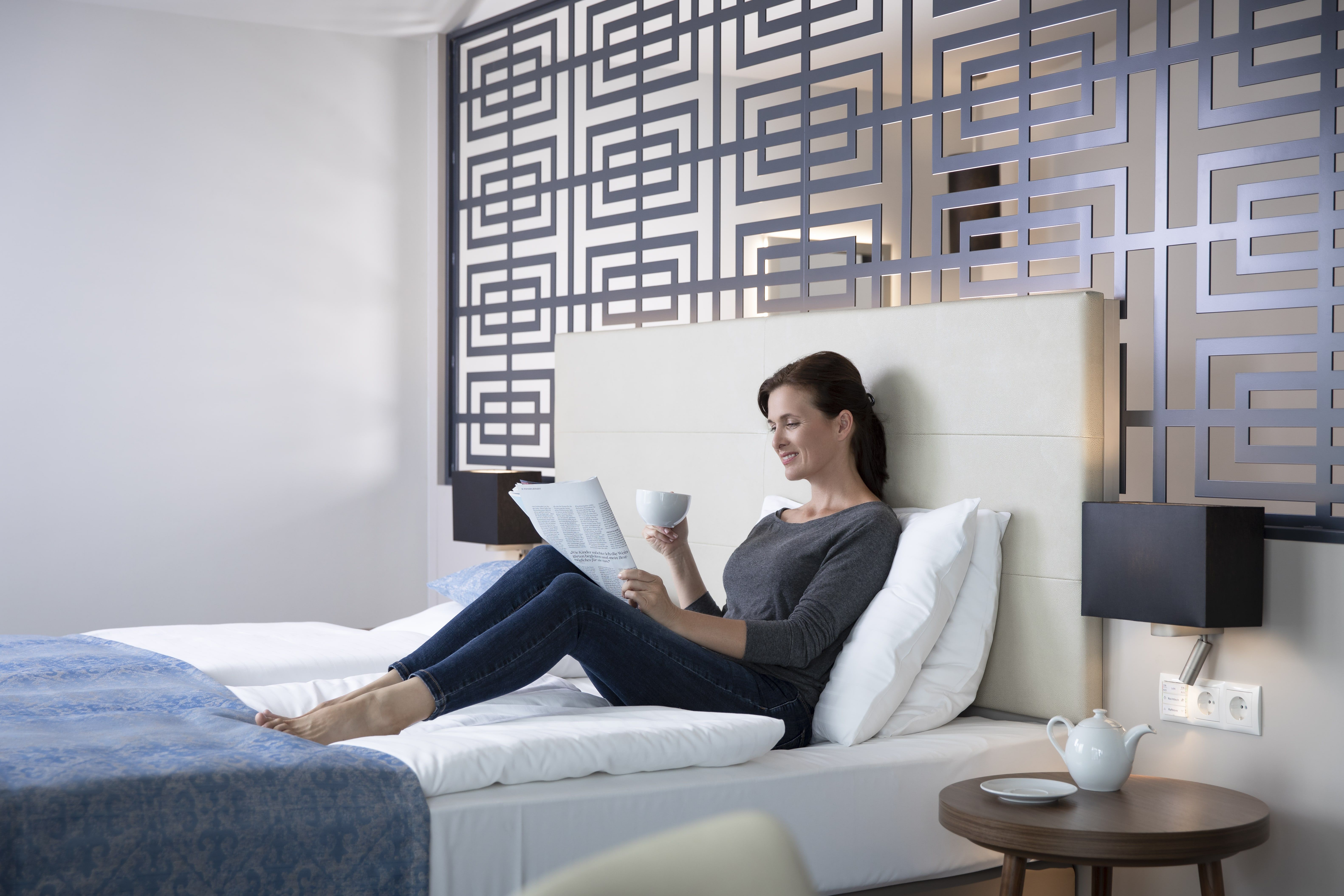 Woman relaxing on a bed in a modern hotel room, reading a newspaper and holding a cup. Decorative wall in the background.