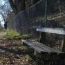 A hiking trail with a wooden bench and a chain-link fence. A person is walking in the distance.