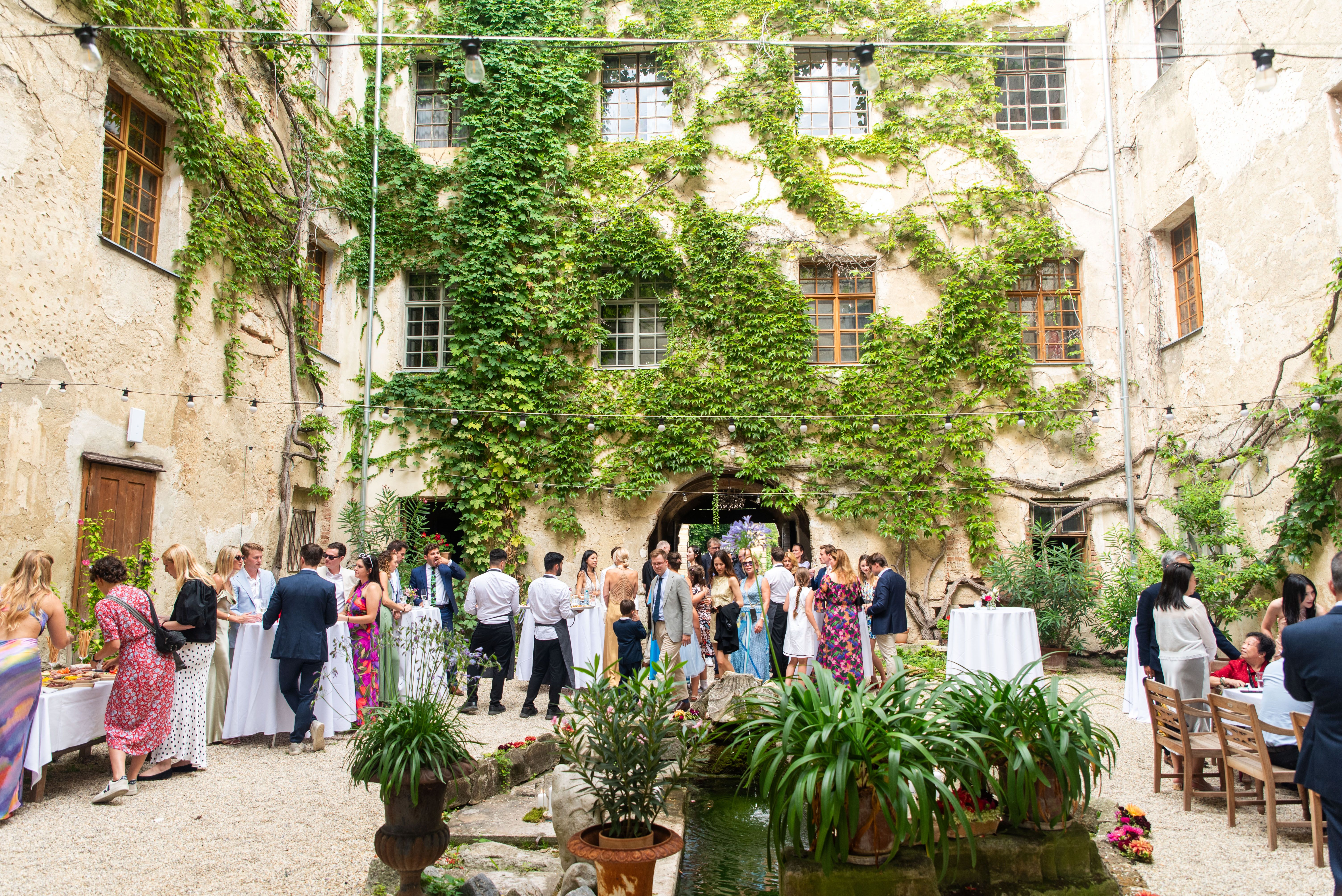 People gather in a leafy courtyard with tables and decorations.