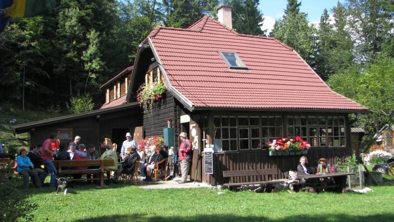 A mountain hut with a red roof, surrounded by forest, with people sitting outside.