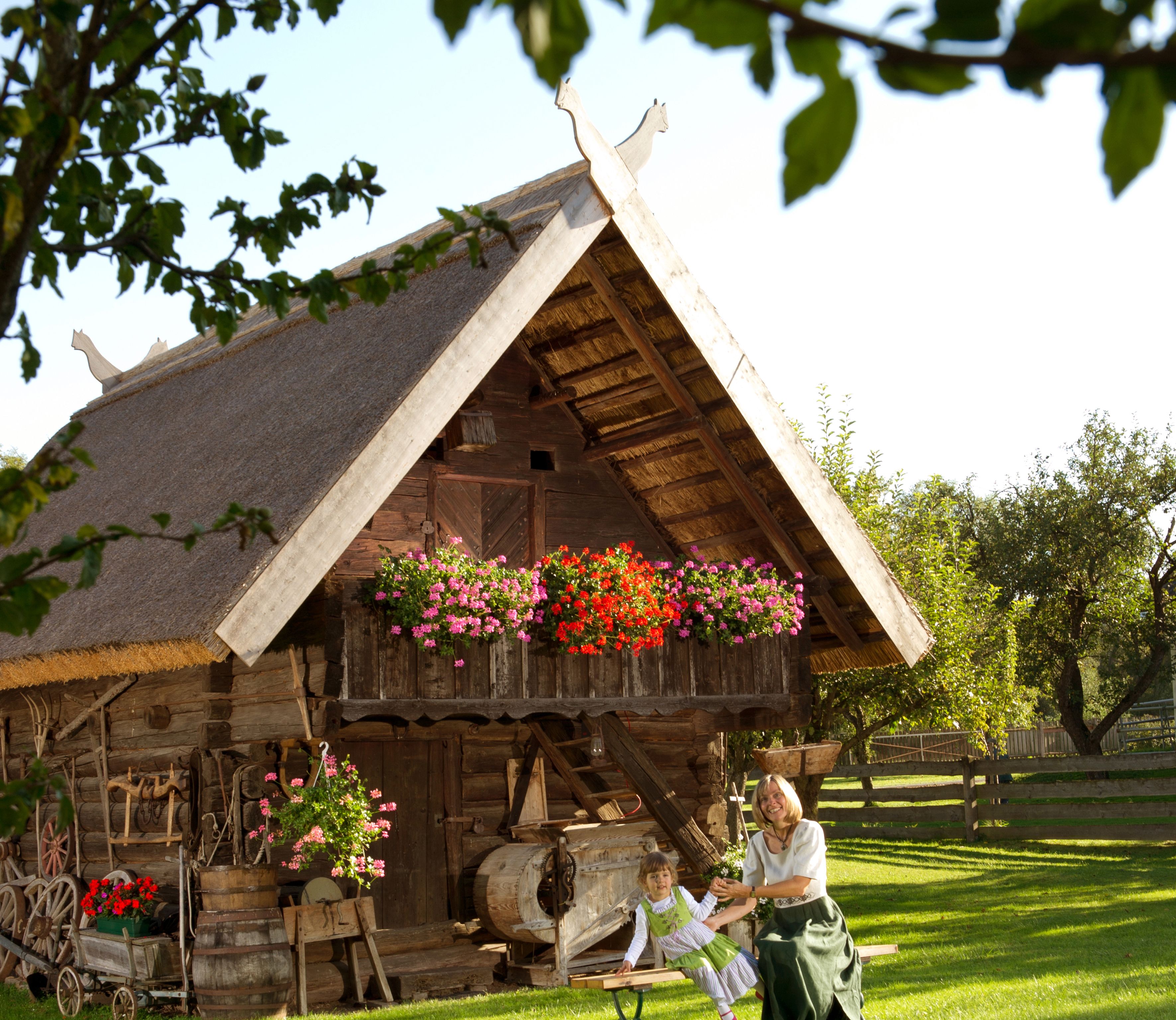 Traditional wooden building with thatched roof and flowers, woman and child in traditional costume in front.