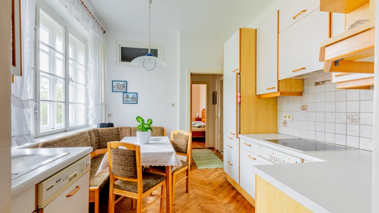 Dining area with wooden furniture, corner bench, table and chairs, bright kitchen with window, plants on the table.