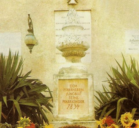 Monument with inscription, surrounded by plants and a basket of potatoes and sunflowers.