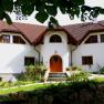 A large, white house with a red tiled roof and a well-tended garden surrounded by trees.